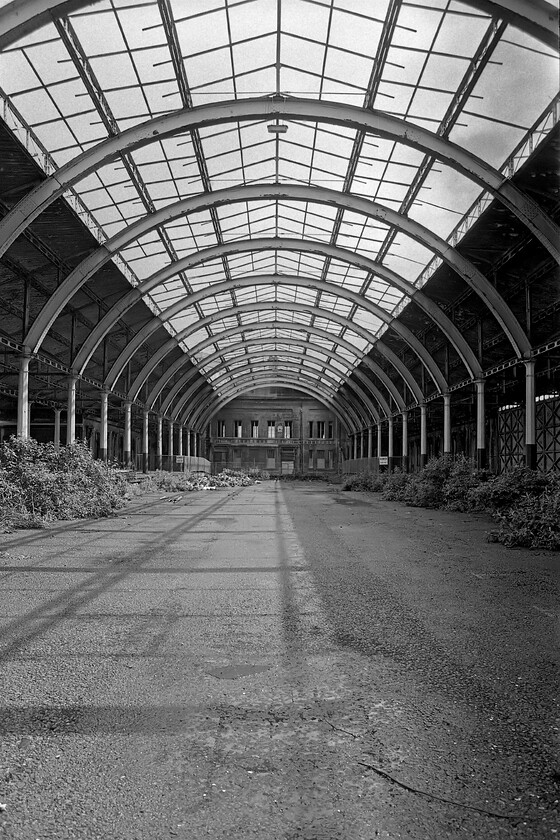 Interior of trainshed, Bath Green Park station 
 Looking down towards the main building at Bath Green Park station reveals that the arched roof has lost all of its glazing. This was not as a result of its closure in 1966 but actually occurred much earlier during the Second World War following bomb damage. In his view, the track beds have been tarmacked over and marked out for car parking. There were four lines with just two platforms. The centre roads were used for the storage of stock. There is a reversed angled view of this one at.... https://www.ontheupfast.com/p/21936chg/30023071330/x11-interior-trainshed-bath-green 
 Keywords: Interior of trainshed Bath Green Park station