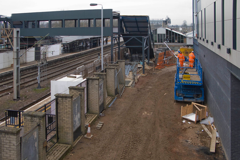 New footbridge & station building, Northampton station 
 With the old station building still functional and in use it is somewhat dwarfed by the new one to the right that is having its cladding attached by the workers in the cherry picker. The new footbridge will join to the station structure at the far end above where the workers are located. The mud-covered road in the foreground used to give access to the station's long stay car park. 
 Keywords: New footbridge station building Northampton station