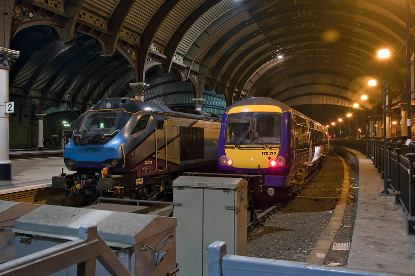 68024 & 170472, stabled, York station 
 A picture that clearly illustrates how different types of lighting are recorded on modern cameras (and silver halide film for that matter). In this picture, taken at York, there is LED lighting to the left in the main train shed that is recorded as a very clean and white light. However, to the right is more traditional sodium illumination that gives the 'warm' orange and hazy lighting effect. Photoshop can correct the colour hue thrown up by different light sources but I have kept this virtually 'as taken' to show the effects. 68024 'Centaur' stands stabled at platform two whilst 170472 does the same next to it. 
 Keywords: 68024 170472 York station