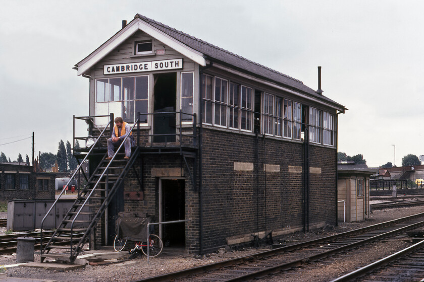 Cambridge South signal box (LNER, 1925) 
 Cambridge South signal box was located on the platform end of the station so was easy to photograph as seen here. It is an LNER box dating from 1925 that has had its locking room windows bricked up probably as a wartime blast protection measure. The box controlled an odd mix of mechanical signals and colour lights using electronic control rather than the more conventional levers. The box was to close on 16.10.82 with control moving to the newly built PSB with electrification arriving six years later. Notice the rail worker taking some sort of break on the box steps, today he would be head to foot in hi-viz and a white helmet back then, just an orange tabard sufficed! 
 Keywords: Cambridge South signal box LNER London and North Eastern Railway