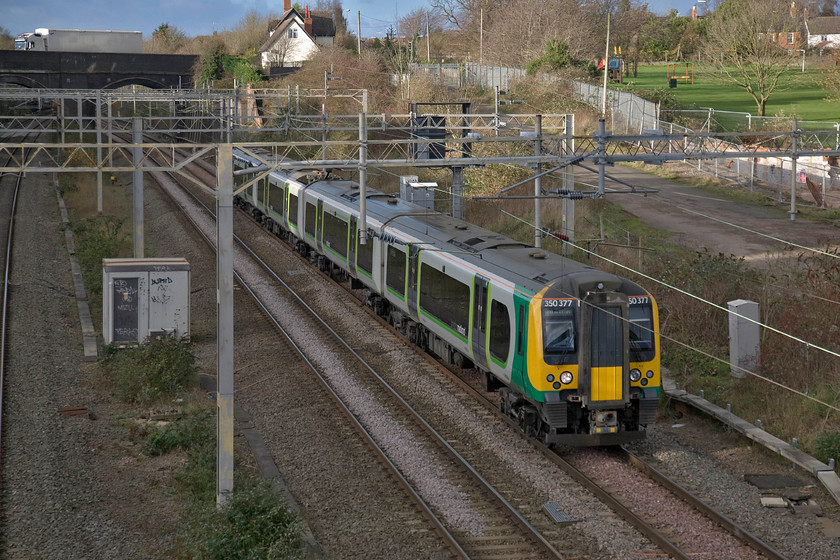 350377, LM 13.14 Birmingham New Street-London Euston (1Y46), site of Road station 
 It's already 14.30 and the sun has dropped so it does not manage to illuminate the 13.14 Birmingham New Sreet to Euston as it passes the site of Roade station woked by 350377 working as a single unit. Notice the line of cottages at the top left if the image, these were built for railway when the line through Roade and the cutting just beyond the bridge were being constructed. The navies put up with primitive conditions in encampments with the railway cottages being the preserve of the senior staff overseeing the work. 
 Keywords: 350377 13.14 Birmingham New Street-London Euston 1Y46 site of Road station