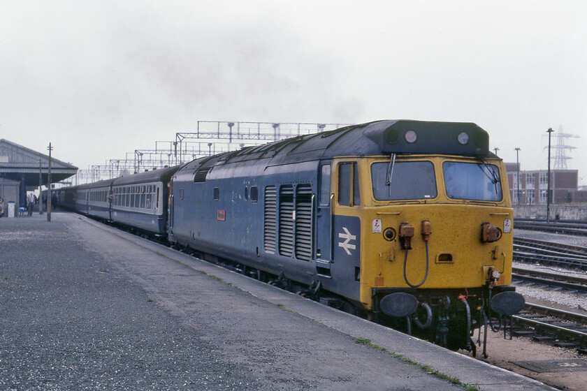 50014, 11.30 London Paddington-Paignton (1B68), Newton Abbot station 
 50014 'Warspite' makes a row as it departs from Newton Abbot station leading the 1B68 11.30 Paddington to Paignton service. The gantries above the stock supported lights that illuminated the extensive carriage sidings in the yard adjacent to the station. The sidings formed part of the relatively new and purpose-built diesel depot constructed at the end of steam ostensively to service the diesel-hydraulic fleets that replaced the Kings, Castles and Halls. The new depot itself did not last very long closing the year that this photograph was taken with all servicing now moving to Laira. A large industrial estate now occupies most of the site. 
 Keywords: 50014 11.30 London Paddington-Paignton 1B68 Newton Abbot station Warspite