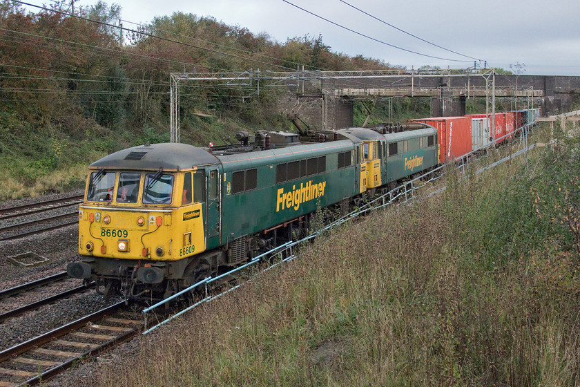 86609 & 86604, 04.57 Trafford Park-Felixstowe North (4L97), Ashton Road bridge 
 Vintage AC power in the form of 86609 and 86604 brings the 04.57 Trafford Park to Felixstowe Freightliner past a new spot created by contractors to Network Rail now that they have completed repairs to the embankment. In all the years that I have lived across the field from this spot, in fact, it's in full view of my kitchen window. It had been heavily wooded and taking photographs of up trains had been impossible. The bridge in the background carries Ashton Road over the WCML just south of Roade. It was constructed in the early 1960s replacing a more traditional bridge in order to accommodate the wiring when the line was in the process of being electrified. 
 Keywords: 86609 86604 04.57 Trafford Park-Felixstowe North 4L97 Ashton Road bridge Roade Freightliner