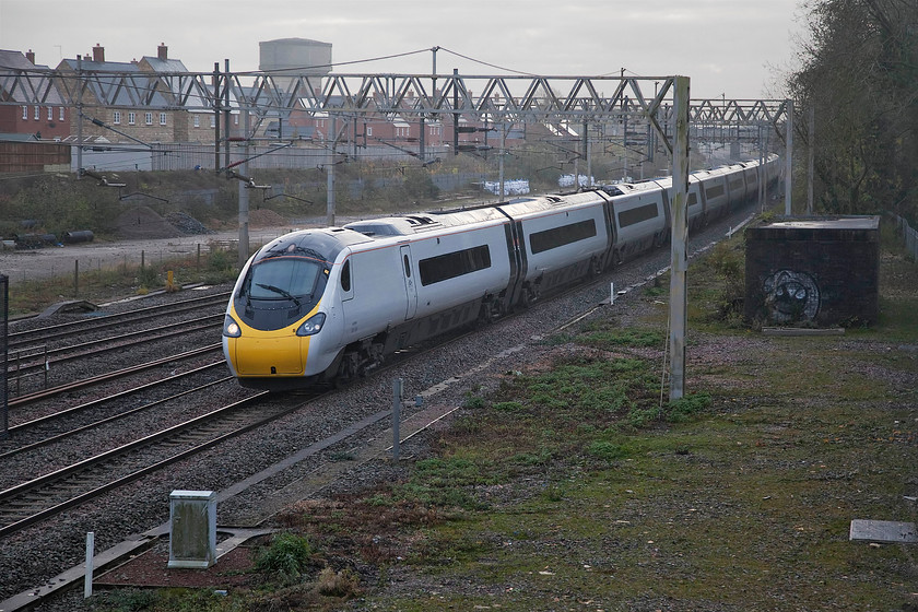 390154, VT 08.20 London Euston-Manchester Piccadilly (1H12, 2L), site of Roade station 
 Looking rather like an Albino train, 390154 heads north with the 08.20 Euston to Manchester Piccadilly. The totally de-branded Pendolino is seen passing the site of Roade station with the vast housing estate in the background that now occupies the site of the former Pianoforte site. This view is a new one as Network Rail has undertaken a huge vegetation clearance exercise in the foreground. However, as it is looking south-east with some tall trees to the right, the photographic opportunities offered by it are a little limited. 
 Keywords: 390154 08.20 London Euston-Manchester Piccadilly 1H12 site of Roade station Pendolino