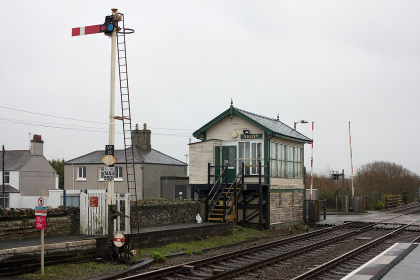 Valley Signal Box (LNWR, 1904) & up starter 
 Valley's up tall up home signal stands at the platform end with the LNWR 1904 signal box behind. Note the round shunting disk signal at the base of the post. This is for the cross-over and spur that links the mainline to the short siding that was used to load and unload the nuclear flasks when Wylfa power station was open. This siding is situated behind the shrubs and trees in the background of the image. 
 Keywords: Valley Signal Box up starter