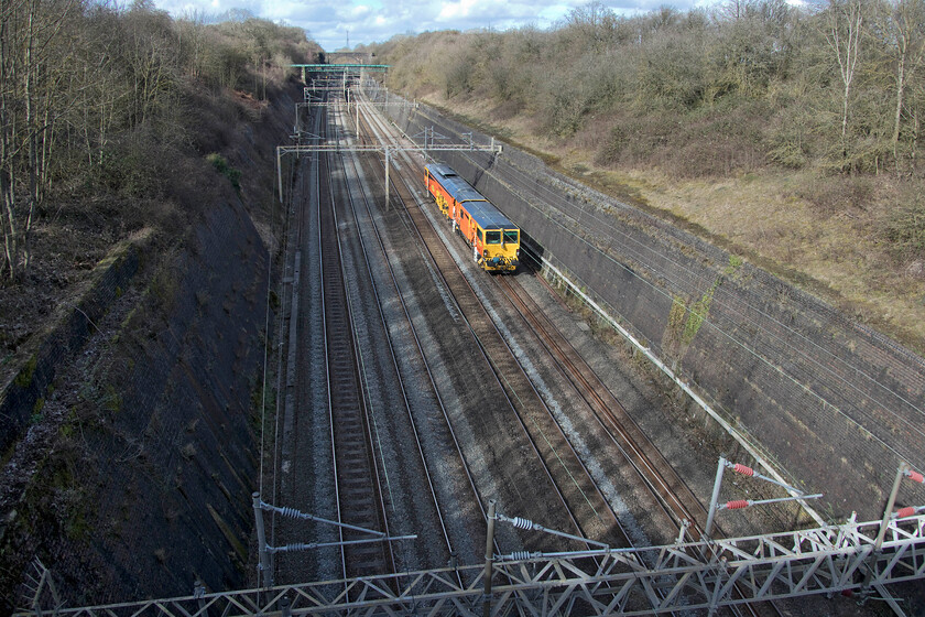 DR73XX, 13.13 Rugby access sidings-Watford London Concrete (35L), Roade cutting 
 For a change, I have adopted a full wide-angled view of Roade cutting taken from one of three bridges that cross it. In the coming year or so a fourth bridge will be constructed between where I am and the green aqueduct in the distance that will carry Roade's much-needed bypass. An unidentified track machine in the DR73XX series heads south through the cutting as the 13.13 Rugby to Watford London Concrete sidings. 
 Keywords: DR73XX 13.13 Rugby access sidings-Watford London Concrete 35L Roade cutting