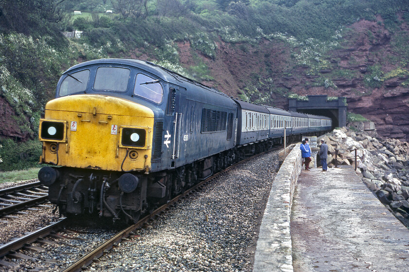 45061, 07.28 Derby-Paignton (1V62), Parson's Tunnel 
 One of my favourite photographs of a Peak! It shows split head coded 45061 emerging from Parson's tunnel leading the 1V62 07.28 Derby to Paignton now covering the final few miles of its journey as it heads along the sea wall towards Teignmouth. The split headcoded examples of the Peaks were a favourite type in my eyes and I was always pleased when I captured one on film including another example (45024) dominating the home page of this website. I have always been curious about the three people standing at the top of the rather slippery and small steps on to the sea wall in this scene, they appeared to be enthusiasts but are taking no notice of the passage of this train probably uninterested due to it being hauled by a rather boring and mundane Peak on some Mk. 1 stock; what they would give for that scene today! 
 Keywords: 45061 07.28 Derby-Paignton 1V62 Parson's Tunnel Peak split headcode