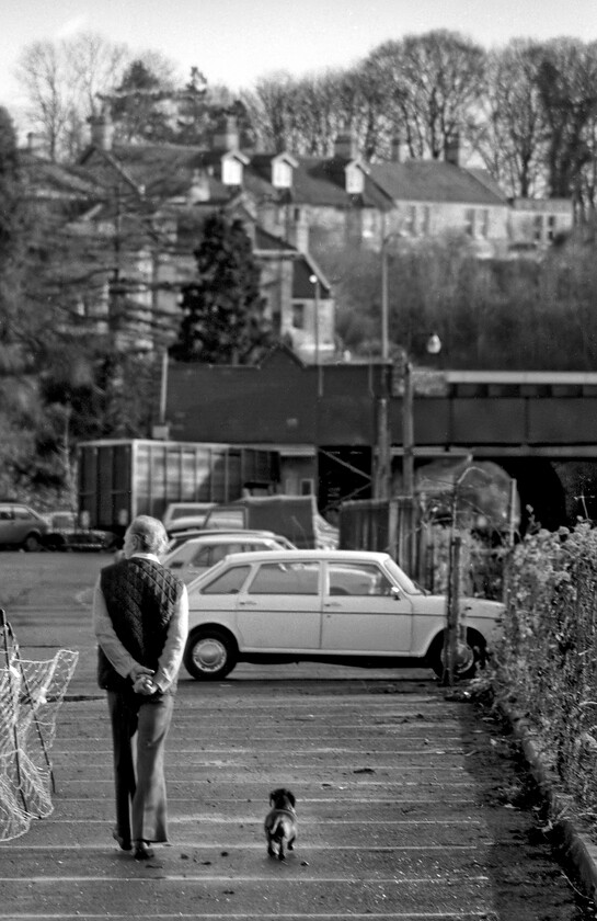 Station car park, Bradford-on-Avon 
 A man walks his dog through Bradfrod-on-Avon's station car park perhaps heading back towards his white Austin Maxi? Bradford's station can be seen in the background with its footbridge and the short tunnel. The car park occupies the site of the former goods yard and goods depot that was located where the line of cars is seen right in front of the camera. 
 Keywords: Station car park Bradford-on-Avon