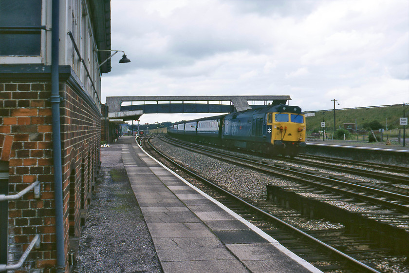 50028, 07.30 London Paddington-Paignton (1B24), Tiverton Junction station 
 On a particularly dull July day, 50028 'Tiger' heads westwards at Tiverton Junction station leading the 1B24 07.30 Paddington to Paignton service. BR was in the throws of running down Tiverton Junction station providing it with a poor service pattern and doing very little to maintain it. Patronage was low, which can be witnessed by the empty station car park behind the down platform. The station was closed on 11.05.86 with the opening of Tiverton Parkway station. 
 Keywords: 50028 07.30 London Paddington-Paignton 1B24 Tiverton Junction station Tiger