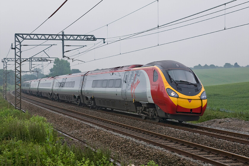 390039, VT 07.20 London Euston-Manchester Piccadilly (1H09), Milton crossing 
 On a particularly grey, dull and chilly June morning 390039 speeds north past Milton crossing just north of Roade cutting that commences in the trees above the rear of the train. The Pendolino is working the 07.20 Euston to Manchester 1H09 Virgin Trains West Coast service that left the capital some thirty-five minutes previously. 
 Keywords: 390039 07.20 London Euston-Manchester Piccadilly 1H09 Milton crossing Virgin Trains West Coast Pendolino