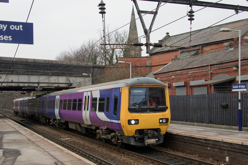 323226, NT 10.59 Manchester Piccadilly-Hadfield (2G11, 1L), Guide Bridge station 
 With the spire of Audenshaw's St. Stephens church above the scene, 323226 arrives at Guide Bridge station working the 10.59 Manchester Piccadilly to Hadfield service. The station enjoys a regular service with trains arriving and departing at frequent intervals. The once extensive yards to the east of the station in which I photographed lines of class 76s when I passed in October 1978, have been removed but the land is still largely extant. Notice in this image the electrical stanchion supporting the catenary. This metalwork is the original installation from when the line was electrified for 1.5 kV DC operation in 1955. It has had the wiring replaced along with all the other infrastructure and now operates at 25 kV AC. 
 Keywords: 323226 10.59 Manchester Piccadilly-Hadfield 2G11 Guide Bridge station