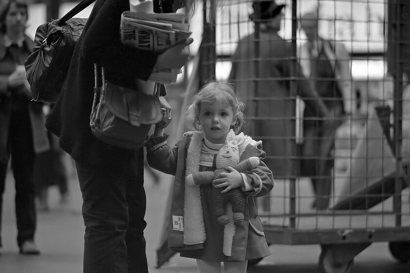 Mother & daughter, London Paddington station 
 A mother and her slightly apprehensive looking daughter wait on Paddington station's busy and noisy concourse preparing to make a journey westward bound. Whilst the little girl clutches her beloved woven companion her mother takes the travelling bag on her shoulder and newspapers for the journey in her arm. I have been able to make out that the newspaper in view is The Mirror and having researched the headline for the day I can state that it reads 'WHAT THE RIPPER TOLD HIS WIFE'. There is also a picture of Peter William Sutcliffe, the Yorkshire Ripper, on his wedding day. This story was being covered by the paper, and every other one for that matter, as his trial for the murder of thirteen women and attacks to a further ten had started the previous day, the verdict is now history off course and I suppose at the time of writing during the COVID pandemic has an added irony as Sutcliffe died from the virus on 13.10.20 having refused prison hospital treatment 
 Keywords: Mother daughter London Paddington station